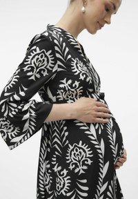 Robe florale noire avec des motifs blancs, présentant des manches cloche 3/4 et un lien à la taille, portée par une personne tenant son ventre rond.
