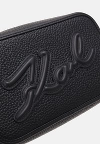 Borsa in pelle nera con superficie texture e ricamo del marchio in rilievo. Forma arrotondata con contorni lisci e dettagli cuciti.
