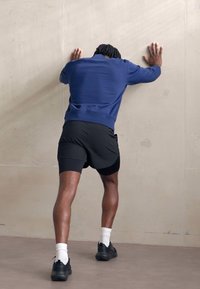 Uomo atletico in felpa blu navy e pantaloni corti neri, appoggiato a un muro chiaro, indossando sneakers nere e calzini bianchi.