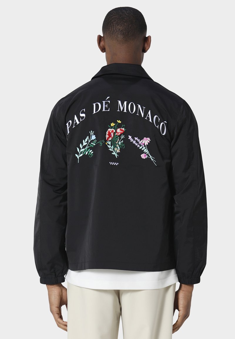 Persoon draagt een zwarte jas met de tekst "Pas Dé Monaco" en kleurrijke bloemenborduursels op de achterkant, gecombineerd met lichtgekleurde broek.