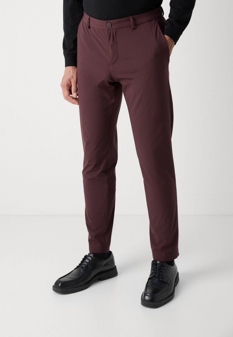 Burgunderfarbene Slim-Fit-Hose aus glattem Stoff, mit Knopfverschluss, Vordertaschen und Bündchen. Kombiniert mit schwarzen Schnürschuhen.