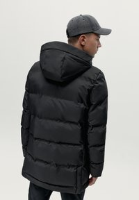 Melns pūkains jaka ar kapuci, ar quilted sekcijām un gludu apdari. Valkāta ar tumši pelēku cepuri, kas demonstrē vienkāršu un modernu dizainu.