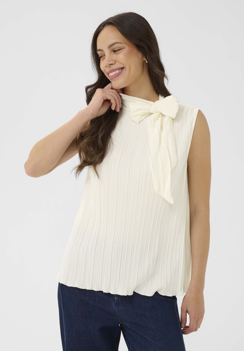 Femme souriante, portant un haut blanc plissé sans manches avec un grand nœud au col et un jean bleu foncé, debout devant un fond blanc uni.