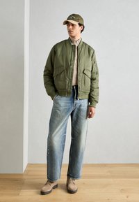 Olivengrønn bomberjakke med to frontlommer, beige genser, lyseblå løse denimjeans og beige støvler på tregulv.