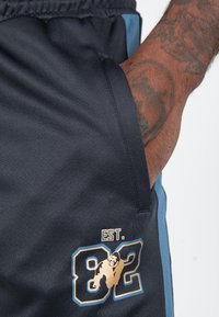 Marineblauwe sportshorts met een blauwe zijpaneel, voorzien van een gouden "EST. 82" grafiek en een gorilladesign. Gelaagd stof en zijzak zichtbaar.