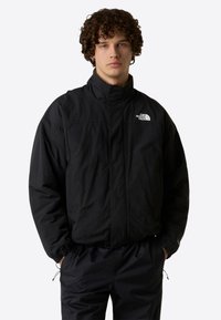 Chaqueta aislante negra con un cuello alto, que presenta un logo blanco en el lado izquierdo del pecho y puños elásticos. Textura suave, diseño práctico.