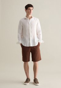 Camicia bianca di lino con maniche lunghe, pantaloni corti di lino marroni e sneaker marroni. Modello in piedi su uno sfondo neutro.