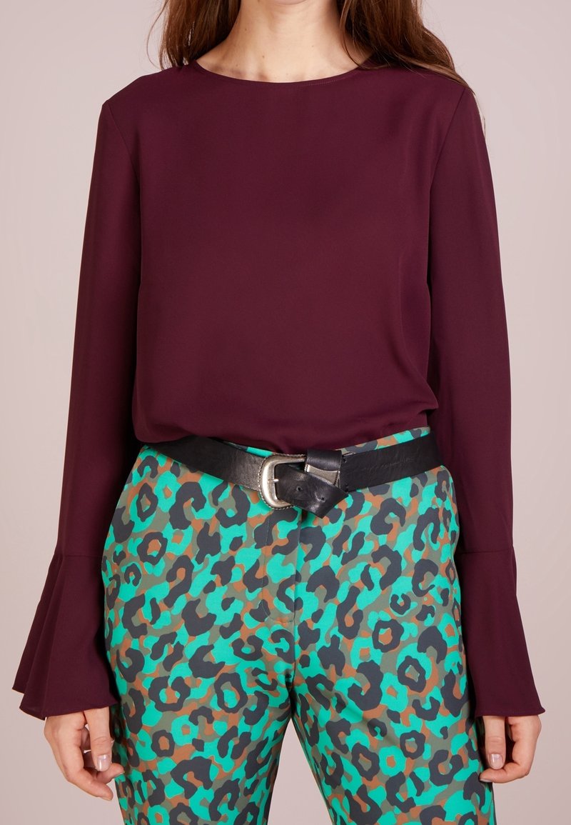 Femme portant un haut bordeaux à manches longues avec des poignets évasés et un pantalon imprimé léopard turquoise, marron et noir avec une ceinture noire.