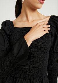 Svart klänning med fyrkantig urringning, rynkad överdel och puffärmar. Slätt tyg med ett mönster av textur som visar detaljerad sömnad.