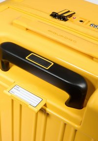 Valigia rigida gialla con scanalature verticali, manico nero, portanome e due cursori neri della cerniera. Logo del marchio visibile in alto.