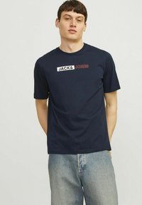 Marineblauwe katoenen t-shirt met een rechthoekig grafisch logo in wit en rood, geplaatst op de bovenste linker borst. Korte mouwen, ronde hals.