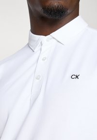 Balta poloshirta izgatavota no kokvilnas, ar īsām piedurknēm, ar pogājamu apkakli un melnu "CK" logo uz krūšu. Tekstūrēts audums.
