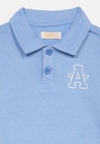 Ανοιχτό μπλε πόλο μπλουζάκι από απαλό βαμβάκι, με γιακά, λοξή σειρά με δύο κουμπιά και λευκό γραφικό γράμμα «A» στο στήθος.