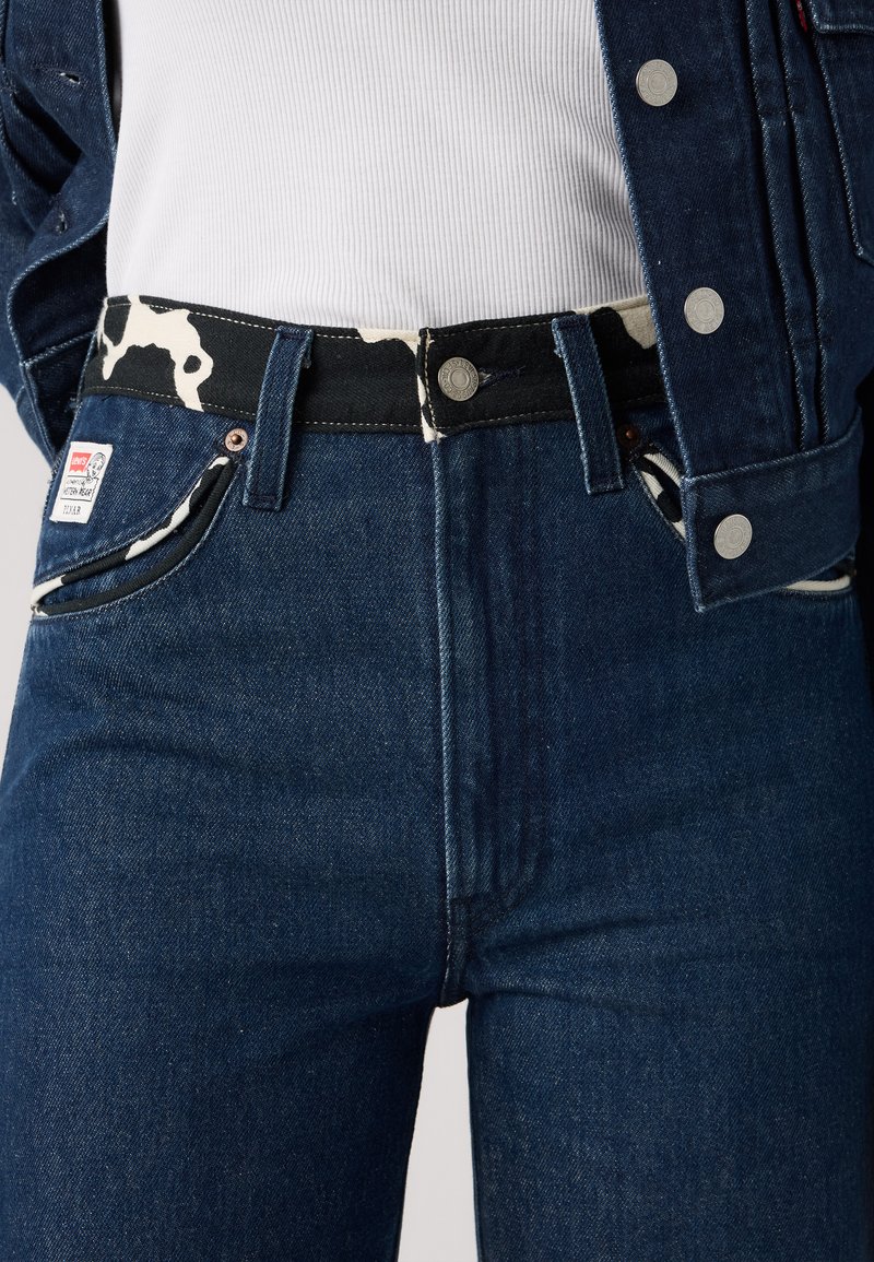 Mørkeblå denimbukser med et sort og hvidt kvægprint i taljen, med metalknapper, frontlommer og et mærke på lommen.