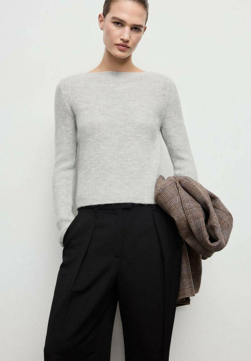 Suéter de canalé gris claro con mangas largas, combinado con pantalones negros con pliegues. Un blazermarrón a cuadros está colgado del brazo.