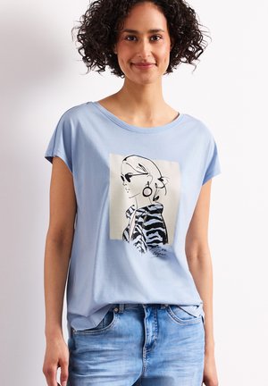 Femme souriante aux cheveux bouclés portant un t-shirt bleu clair avec une illustration en noir et blanc d'une femme élégante et un jean bleu.