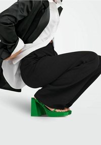 Sandales vertes à talons épais avec une base large et des fines brides, associées à un pantalon noir à jambes larges et une chemise blanche sous un blazer noir.