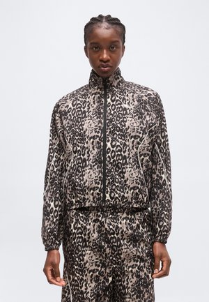 Personne aux cheveux tressés portant une veste zippée et un pantalon assortis à imprimé léopard, debout devant un fond clair uni.