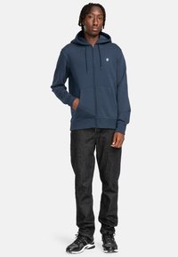 Marineblaue Zipperjacke mit Kängurutasche und Kapuze mit Zugband, aus weichem Material gefertigt. Kombiniert mit dunklen Jeans und schwarzen Sportschuhen.