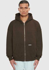 Hoodie zippé marron avec capuche, grandes poches avant et poignets côtelés. Fabriqué en tissu doux avec un éclat subtil. Étiquette sur la poche inférieure.