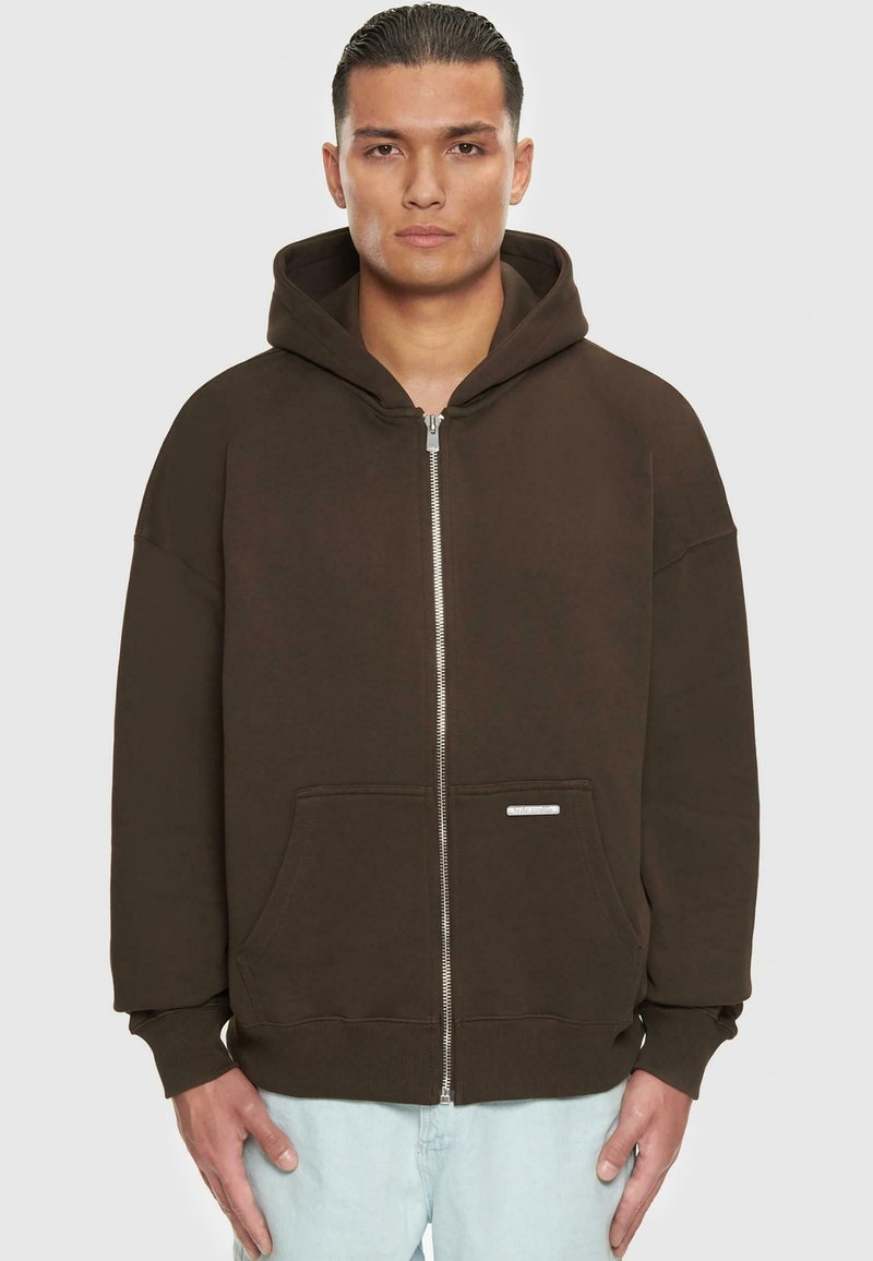 Hoodie zippé marron avec capuche, grandes poches avant et poignets côtelés. Fabriqué en tissu doux avec un éclat subtil. Étiquette sur la poche inférieure.