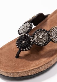 Sandalo con fascia in perline che presenta decorazioni circolari nere, argento e grigie su una soletta in suede marrone con una suola nera testurizzata.