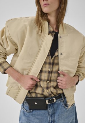Woman adjusting beige bomber jacket over plaid shirt with black undershirt, wearing blue jeans and black leather belt pouch.
