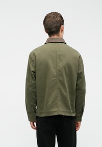 Maslinasto zelena jakna s smešnim kordura ovratnikom, teksturirana tkanina i opušteni kroj. Vidljive šavove i detalj na leđima. Crne hlače.