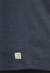 Dunkelblaues T-Shirt aus strukturiertem Stoff, mit einer genähten Naht und einem rechteckigen weißen Markenlabel in der linken unteren Ecke.