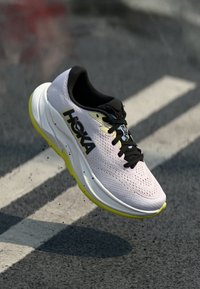 Running shoe featuring a light pink upper with ventilation holes, black laces, and a yellow sole. "HOKA" logo prominently displayed on the side.