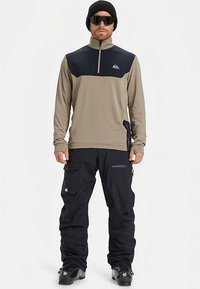 Uomo con berretto nero, grandi occhiali da sole, giacca zip beige e nera, pantaloni cargo neri e stivali da sci neri, in piedi di fronte.