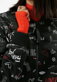 Giacca bomber nera con grafiche ricamate in rosso e bianco. Caratterizzata da polsini neri a coste e un maglione a collo alto rosso brillante indossato sotto.