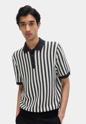 Junger Mann mit kurzen dunklen Haaren, der ein schwarz-weiß gemustertes kurzärmeliges Poloshirt trägt, die Hände in den Taschen, vor schlichtem Hintergrund.