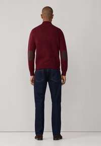 Maglione bordeaux con colletto alto, dotato di toppe marroni sui gomiti, abbinato a jeans blu scuro. La trama appare morbida e lavorata a maglia.