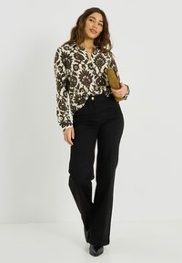 Blusa de estampado floral en crema y marrón con cuello, combinada con pantalones negros de cintura alta y tacones negros, sosteniendo un bolso de mano.