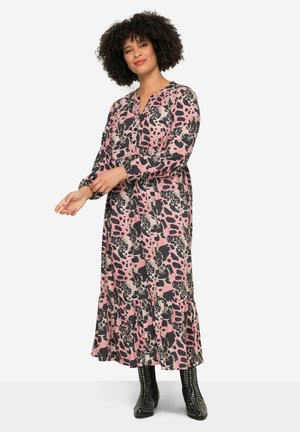 Robe longue, rose et noir à motifs, avec un ourlet froncé et des manches longues, fabriquée en tissu léger. Modèle portant des bottines à talons noires ornées de clous.