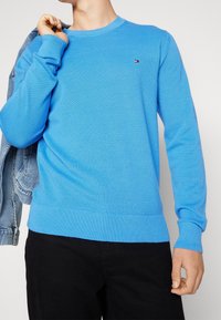 Maglione a coste blu con maniche lunghe e scollatura a girocollo. Presenta un piccolo logo sul petto. Indossato con una giacca di jeans appoggiata sul braccio sinistro.