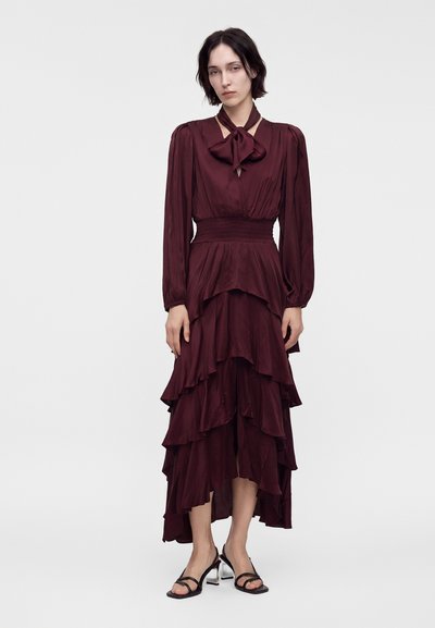 Femme portant une longue robe bordeaux à volants superposés, manches longues, taille haute et encolure à nœud papillon, associée à des sandales à talons noires.