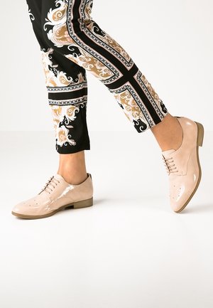 Chaussures beiges en cuir verni à lacets avec un design brogue, dotées d'un bout rond et d'un petit talon en bois, associées à un pantalon noir à motifs.