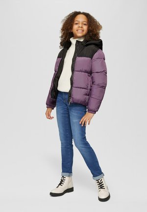 Veste matelassée violette et noire avec une capuche, fermeture éclair sur le devant et poignets élastiques, portée sur un pull crème et un jean bleu, associée à des bottes blanches.