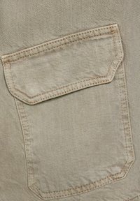 Poche en tissu beige avec un rabat et des détails de couture visibles, probablement partie d'un vêtement tel qu'un pantalon ou une veste.