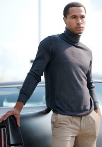 Dark gray turtleneck sweater in a soft knit, fitted design, with long sleeves, paired with light beige pants. Background includes a classic car.