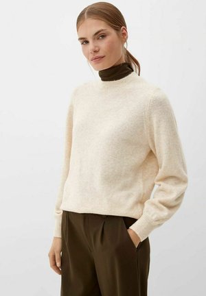 Frau mit leicht beigem Pullover über einem dunkelbraunen Rollkragenpullover, dunkelbraunen Hosen, die mit einer Hand in der Tasche vor einem schlichten Hintergrund steht.