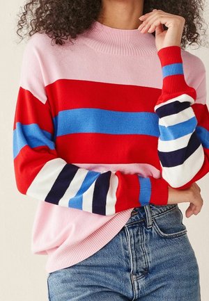 Femme portant un pull rose avec des rayures horizontales rouges, bleues, blanches et bleu marine, associée à un jean en denim bleu, les bras croisés.
