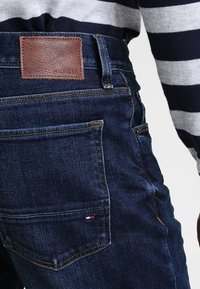 Tmavě modré džíny z denimu s koženou značkovou nášivkou se štítkem "HILFIGER" na zadní straně. Mají klasický střih a několik kapes.