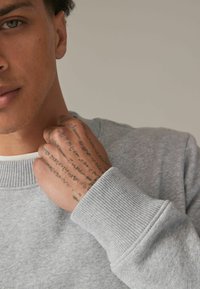 Lichtgrijze sweater met ribgebreide manchetten en halslijn. Close-up van een hand met ingewikkelde tatoeages, die lichtjes op het shirt rust. Soepele textuur.