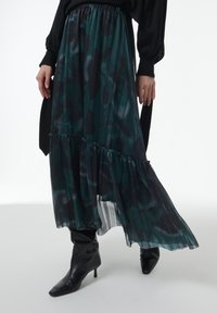 Maxi rok in donkergroen met zwart bloemenpatroon, doorschijnende textuur, geplooid ontwerp en gerimpelde zoom, gecombineerd met zwarte puntige enkellaarsjes.