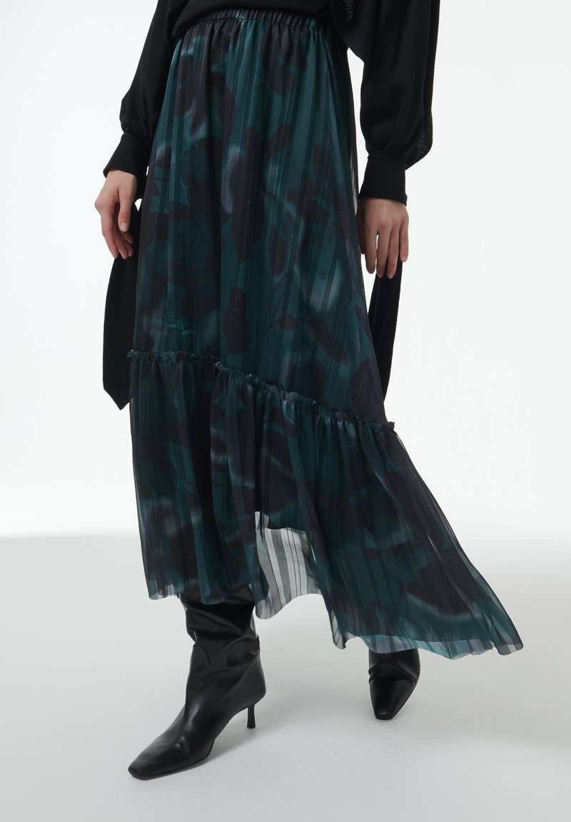 Maxi rok in donkergroen met zwart bloemenpatroon, doorschijnende textuur, geplooid ontwerp en gerimpelde zoom, gecombineerd met zwarte puntige enkellaarsjes.