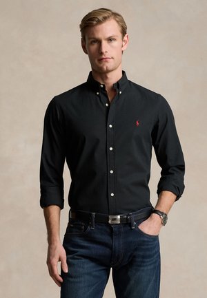 Homme portant une chemise noire à manches longues avec un petit logo rouge, un jean foncé et une ceinture noire, debout avec une main dans la poche.