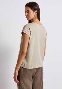Béžové bavlněné tričko s ohrnutými rukávy a uvolněným střihem, v kombinaci se světlými hnědými lněnými kalhotami. Hladká textilie, jednoduchý design, žádné vzory.